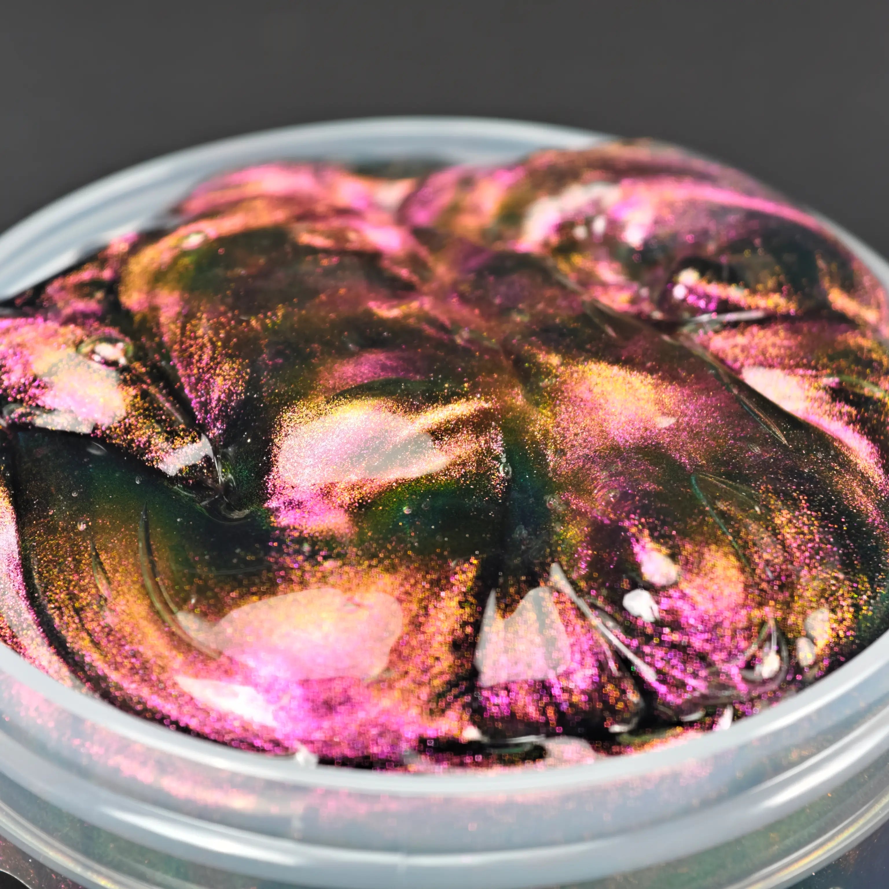 Dark holographic color-shifting slime in a clear jar, featuring metallic gold, pink, and green pigments. Coated putty texture slime for sensory play and stress relief