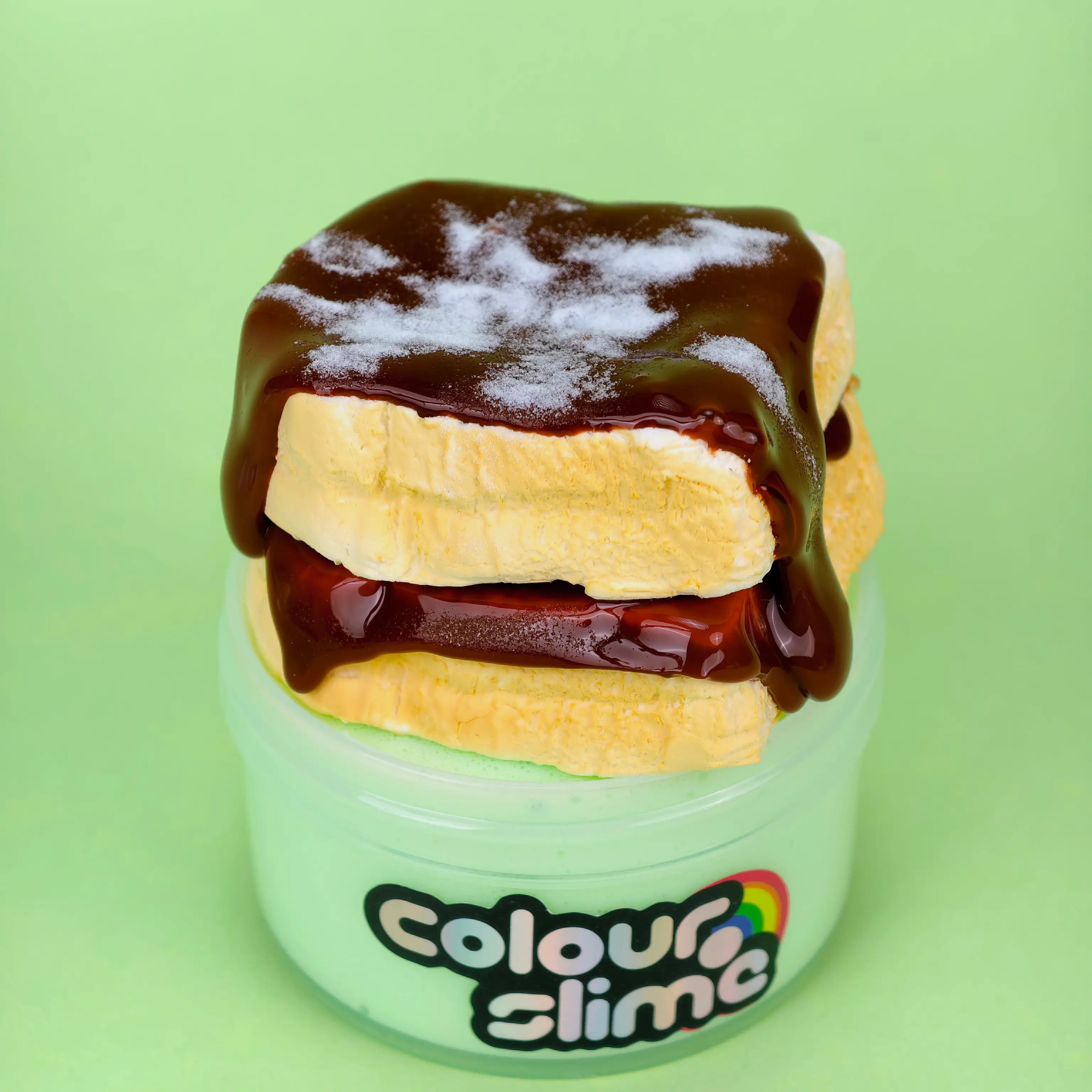 DIY slime kit named Sunday Morning Stack featuring an avocado green base in a jar. It is topped with two realistic air-dry clay toast slices, drizzled with glossy brown chocolate-colored slime, and sprinkled with faux powdered sugar